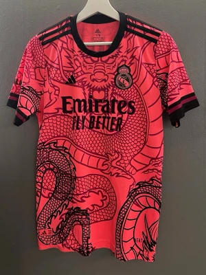 Real Madrid matchtröja  - Säljer en rosa Real Madrid matchtröja från Adidas med svart drakmönster och svarta detaljer. Tröjan har korta ärmar, rund hals och tryck med 'Emirates Fly Better' på framsidan samt 'Mbappé 9' på ryggen. Perfekt för dig som älskar fotboll och unika matchtröjor.