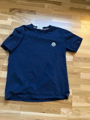 Mörkblå t-shirt med logga - En stilren mörkblå t-shirt i bomull med rund hals och korta ärmar. På bröstet sitter en liten broderad logga som ger en clean och exklusiv känsla. Perfekt till jeans eller shorts för en enkel men snygg look.