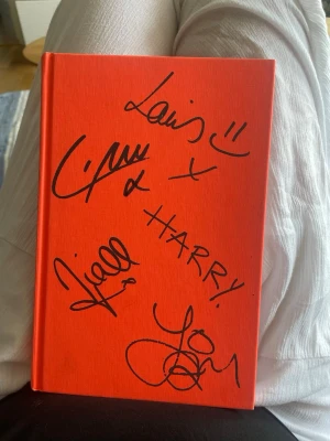 One Direction who we are bok - Säljer en orange hårdinbunden bok med titeln 'Who We Are'. Omslaget är dekorerat med autografer i svart bläck från medlemmarna. Perfekt för fans som vill ha något unikt i bokhyllan. (Inte äkta autografer)