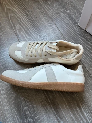 Maison marginella gats  - Stilrena sneakers i vitt och beige med klassisk låg profil. Ovandelen är i en mix av slätt läder och mocka, med beige detaljer och matchande snören. Yttersulan är i gummi med en ljusbrun ton som ger en retrovibe. Perfekta för en clean och avslappnad look. 10/10 skick och riktigt feta 
