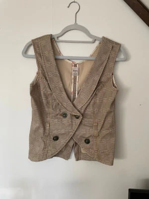 Rutig väst från ONLY  - Snygg rutig väst från ONLY i beige och bruna toner. Västen har bred krage, dubbelknäppning med fyra knappar och ett blommigt innerfoder. Tillverkad i 95% polyester och 5% elastan. Perfekt för att lyfta din outfit med en vintagekänsla.