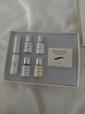 Brow lift kit med borste och vätskor - Komplett brow lift kit från IbcccnDC med fyra små flaskor (cleanser, perm, fixation, nutrition), en vit tub och en brow brush. Perfekt för att forma och vårda ögonbryn hemma. Förpackningen är vit och stilren.