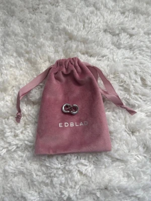 Smycke från Edblad i silverfärg - Säljer ett stilrent smycke från Edblad i silverfärgad metall. Smycket har två sammanlänkade ringar och kommer i en rosa sammetsliknande påse med band. Perfekt accessoar för dig som gillar minimalistisk design.