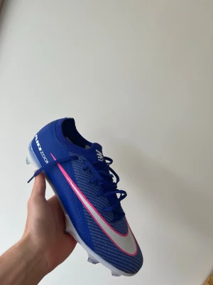 Blå fotbollskor  - Snygga  mörkblåa, nästan lila  fotbollsskor med vit och rosa swoosh längs sidan. Skon har snörning, låg profil och är tillverkad i ett lätt syntetmaterial för bästa bollkontroll och fart på planen. Perfekt för dig som vill sticka ut på matchen. Endast testade av mig på planen. Säljer eftersom jag fick andra fotbollskor istället.