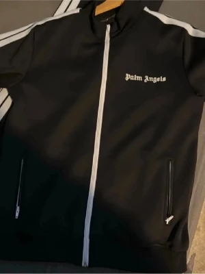 Svart track jacket från Palm Angels - Snygg svart track jacket från Palm Angels med vita detaljer och dragkedja framtill. Jackan har vita ränder längs ärmarna, ståkrage och två fickor med dragkedja. Perfekt för en sportig och trendig look.