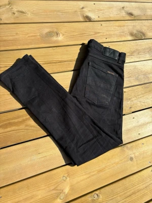  Nudie Jeans Lean Dean - Svarta jeans från Nudie Jeans i modellen 31/30. Jeansen har klassisk femficksdesign, svarta sömmar och Nudie Jeans logga på bakfickan. Tillverkade i ekologisk bomull med en straight passform som sitter snyggt. Perfekta för dig som gillar stilrena och enkla jeans.