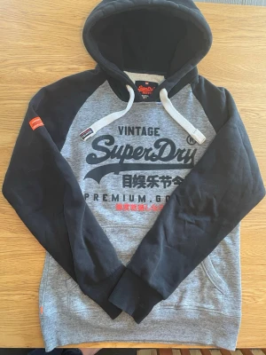 Vintage Superdry hoodie Str L - Snygg hoodie från Superdry i storlek L. Tröjan är grå med svarta ärmar och huva, har vit dragsko och tryck på bröstet i svart och rött. Klassisk känguruficka framtill och ribbade muddar. Perfekt för en avslappnad streetwear-look.