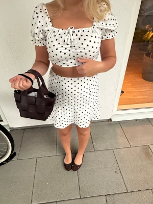 Vit polka dot sett  - Söt vit /svartprickigt sett. kortkjol med svart prickigt mönster. Kjolen har en lätt utställd form och är perfekt för varma dagar. Matchande crop top för en trendig look.