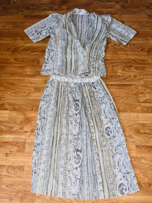 Mönstrat set med kjol och topp - Säljer ett tvådelat set med kjol och kortärmad topp i ljusa toner av beige, grått och vitt. Setet har ett retroinspirerat mönster med blommor och ränder. Kjolen är midilång och toppen har v-ringning och krage och pryds med knappar. Perfekt för dig som gillar vintage vibes!