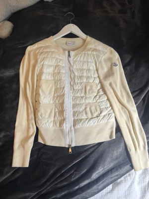 Moncler cardigan - Snygg vit cardiganjacka från Moncler med quiltad front och två fickor. Jackan har ribbstickade muddar, rund halsringning och dragkedja framtill. Ärmarna är i mjuk stickad bomull och loggan sitter på vänster ärm. Perfekt för lager på lager.