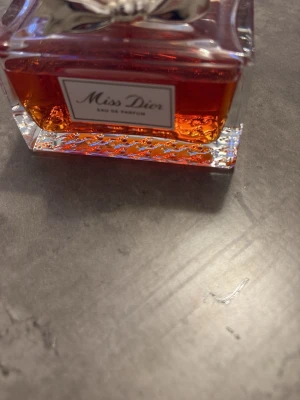 Miss Dior Eau de Parfum - Miss Dior Eau de Parfum i en elegant, rektangulär glasflaska med silverfärgad rosett och detaljerad botten. Parfymen har en varm, bärnstensfärgad ton och flaskan har en exklusiv känsla med tydlig Dior-logga på korken.
