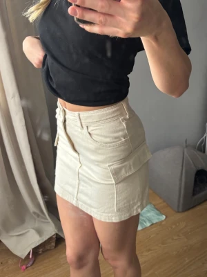 Beige kortkjol med fickor - Snygg beige kortkjol i bomull med sidofickor och knapp- och dragkedjestängning framtill. Kjolen har en rak passform, enkel men trendig. 