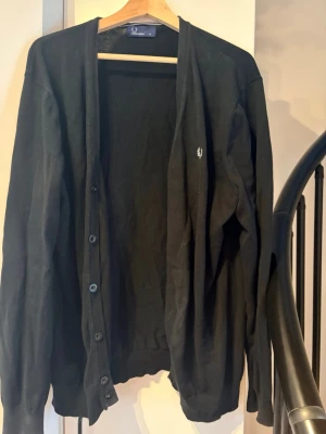 Svart cardigan från Fred Perry XL - Klassisk svart cardigan från Fred Perry med knappstängning framtill och diskret logga broderad på bröstet. Tröjan har långa ärmar och ribbade muddar vid ärmslut och nederkant. Perfekt att slänga över en t-shirt för en clean look.