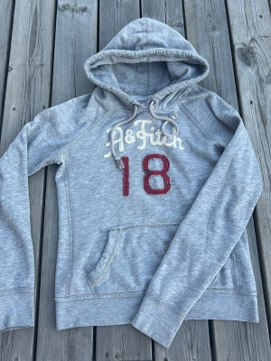 Grå hoodie med tryck från A&Fitch - Snygg grå hoodie med huva och dragsko från A&Fitch. Framsidan har broderad text och siffran 18 i rött. Klassisk känguruficka och mjukt material som känns skönt mot huden. Perfekt för en avslappnad och sportig stil.
