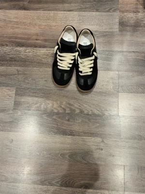 Maison margiela - Snygga svarta sneakers med vita snören och brun gummisula. Skorna har en klassisk låg profil och ovandel i en mix av mocka och slätt material. Perfekta för en stilren och avslappnad look.