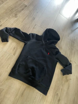 Svart hoodie från Polo Ralph Lauren - Svart hoodie från Polo Ralph Lauren med klassisk känguruficka och röd broderad logga på bröstet. Tillverkad i mjukt bomullsmaterial och har ribbade muddar vid ärmslut och nederkant. Perfekt för en avslappnad och stilren look.