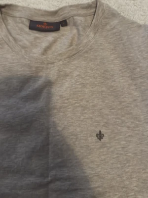 Beige basic t-shirt med liten logga - En enkel grå t-shirt i bomull med rund hals och korta ärmar. På bröstet finns en liten broderad logga i svart som ger en clean och stilren look. Perfekt till jeans eller shorts för en avslappnad vibe.