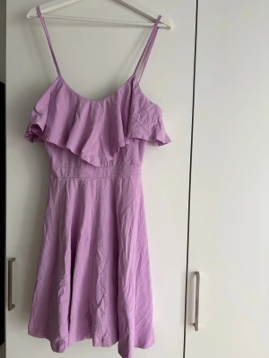 Ljuslila klänning med volang - Söt ljuslila klänning med smala axelband och volangdetalj över bysten. Klänningen har en utställd kjol och är tillverkad i ett lätt tyg, perfekt för varma dagar. Enkel och stilren design som ger en romantisk känsla.