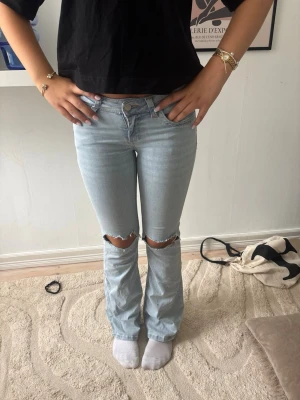 Ljusblå bootcut jeans med hål - Säljer ett par ljusblå bootcut jeans med  hål över knäna och broderade detaljer i rosa på bakfickorna. Jeansen har låg midja och klassisk femficksdesign. Perfekta för en avslappnad och trendig look.