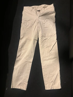Beige raka chinos Oscar jacobsson - Snygga beige chinos med rak passform och klassisk design. Byxorna har framfickor, bälteshällor och två bakfickor med knapp. Tillverkade i ett mjukt bomullstyg som känns bekvämt hela dagen. Perfekta för en clean och stilren look.