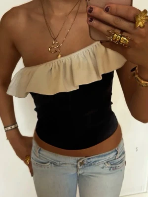 Svart oneshoulder topp med volang - Säljer en svart oneshoulder topp med bred beige volang längs halsringningen. Toppen är tight och croppad, perfekt för dig som vill sticka ut med en unik och trendig look. Passar till både jeans och kjol för en cool outfit.