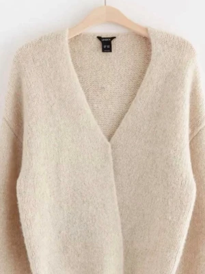 Beige stickad kofta med v-ringning - Mysig beige stickad kofta med djup v-ringning och avslappnad passform. Koftan har långa ärmar och är tillverkad i ett mjukt, fluffigt material som känns skönt mot huden. Perfekt att slänga på sig när det är kyligt ute.