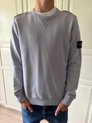 Ljusgrå sweatshirt från Stone Island - Ljusgrå sweatshirt från Stone Island med klassisk rund halsringning och ribbade muddar. Tröjan har den ikoniska svarta logotypen med kompass på vänster ärm. Tillverkad i mjukt bomullsmaterial och har en avslappnad passform. Tröjan är i storlek L men passar mer åt M. Hör av er vid frågor och priset är inte hugget i sten!