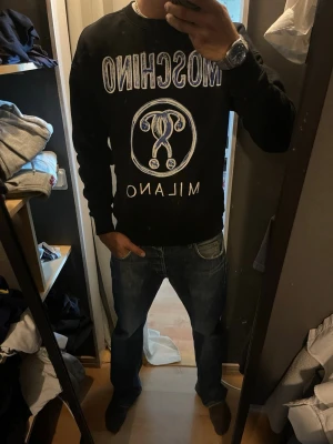 Svart Moschino hoodie med tryck - Jag är 186cm på bilden Säljer en svart hoodie från Moschino med stort vitt tryck på bröstet där det står 'MOSCHINO MILANO' och en grafisk logga i mitten. Hoodien har en klassisk passform och långa ärmar. Perfekt för dig som gillar streetwear och vill sticka ut.