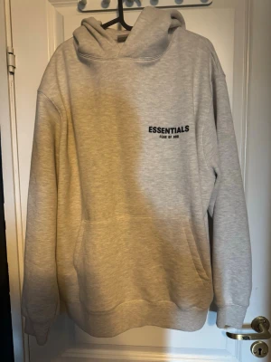 Grå Essentials Fear of God set - Snygg grå hoodie från Essentials Fear of God med svart logga tryckt på bröstet och ryggen. Hoodien har en stor huva, magficka och ribbade muddar vid ärmar och nederkant. Perfekt för en avslappnad och trendig stil.