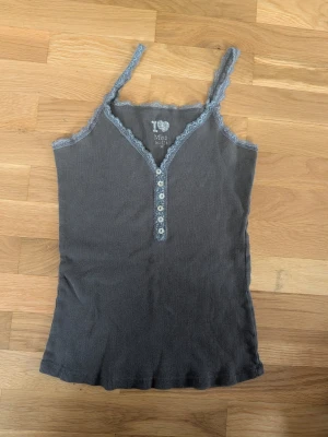 Grå/grön ribbad linnetopp med spets - Snygg grå/grön ribbad linnetopp med spetsdetaljer längs kanterna och v-ringning. Dekorativa knappar framtill ger en extra touch. Perfekt att styla med jeans eller kjol för en trendig look.