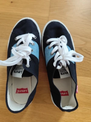 Levi's blå sneakers med vita snören - Snygga sneakers från Levi's i mörkblått tyg med ljusblå denimdetaljer på sidorna. Skorna har vita snören och vita sulor samt klassisk Levi's-logga på insidan. Perfekta för en avslappnad och trendig stil.