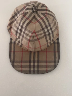 Beige rutig keps från Burberry - Snygg keps från Burberry i klassiskt beige rutigt mönster med inslag av svart, vitt och rött. Justerbar rem baktill med metallspänne. Tillverkad i bomull och har en böjd skärm. Perfekt accessoar för en trendig och tidlös look.