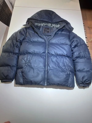 Tommy hilfiger puffer jacket - Snygg mörkblå dunjacka från Tommy Hilfiger med huva och dragkedja framtill. Köpt för 2200kr och har ett par få hål som gör att sy, inga synliga hål!