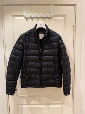 Svart dunjacka från Moncler - Snygg svart dunjacka från Moncler med quiltad design och diskret logotyp på ärmen. Jackan har dragkedja framtill, två sidofickor med dragkedja och en ficka på ärmen. Lätt och smidig modell utan huva, perfekt för kyliga dagar. Storlek Large