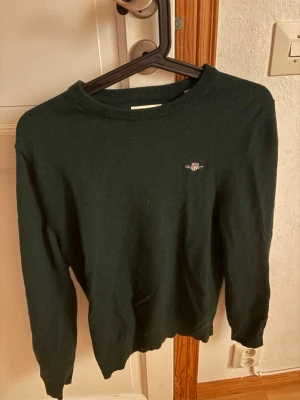 Mörkgrön stickad tröja från GANT - Mörkgrön stickad tröja från GANT i extra fin lammull. Klassisk rund halsringning och diskret GANT-logga broderad på bröstet. Perfekt för dig som gillar stilrena och tidlösa plagg med hög komfort.