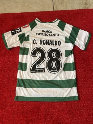 Grönvit Sporting Lissabon tröja - Säljer en klassisk grönvit randig fotbollströja från Sporting Lissabon med C. Ronaldo och nummer 28 på ryggen. Tröjan har klubbmärke på bröstet, sponsortryck och korta ärmar. Perfekt för dig som älskar fotboll och vill sticka ut på planen.