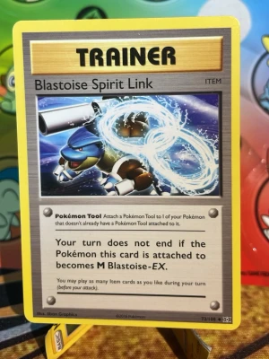 Pokémon Blastoise Spirit Link kort - Pokémonkortet Blastoise Spirit Link är ett tränarkort från Pokémon TCG. Kortet har en färgstark design med Blastoise och vatteneffekter på framsidan. Baksidan har den klassiska blå Pokémon-designen med en Pokéball i mitten.