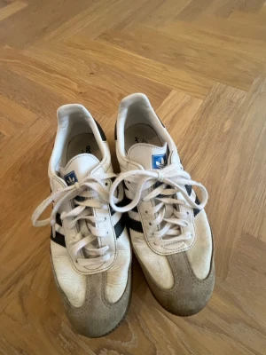 Adidas vita samba sneakers med svarta detaljer - Klassiska Adidas sneakers i vitt läder med svarta ränder på sidorna och grå mockadetaljer vid tån. Skorna har vita snören, blå Adidas-logga på plösen och svart hälparti. Perfekta för en sportig och avslappnad stil. Liten i storlek.