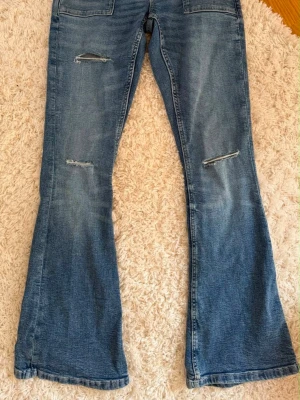 Blå bootcut jeans med slitningar - Säljer ett par blå bootcut jeans med slitningar över knäna och klassiska bakfickor. Jeansen har en lätt utsvängd passform och är gjorda i denim. Perfekta för dig som gillar en avslappnad och trendig stil.