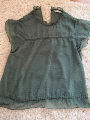 Grön blus med volangärm - Söt grön blus i tunn, transparent chiffong med diskret prickmönster. Blusen har korta volangärmar och liten volang vid halsringningen samt en dekorativ öppning bak. Perfekt att styla med jeans eller kjol för en trendig look.