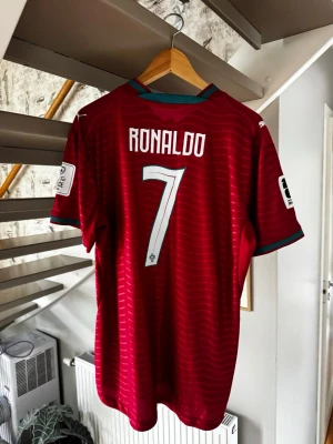Portugal Ronaldo matchtröja röd - Säljer en röd Portugal matchtröja med Ronaldo och nummer 7 på ryggen. Tröjan har gröna detaljer vid krage och ärmslut, samt vita logotyper och landslagsemblem på bröstet. Perfekt för dig som älskar fotboll och vill stötta Portugal.