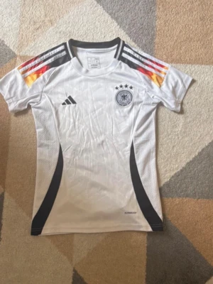 Tysklands  Adidas t-shirt - Vit kortärmad t-shirt från Adidas med Tysklands landslagstryck och fyra stjärnor på bröstet. Svarta och röda detaljer samt gula inslag på axlarna. Tillverkad i polyester och har en sportig, slim passform.