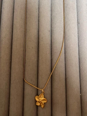 Guldigt halsband med blomma - Snyggt halsband i guldfärg med en detaljrik blomma som hänge. Kedjan är tunn och elegant, och blomman har en fin struktur som ger ett unikt intryck. Perfekt för dig som gillar stilrena accessoarer med en twist.