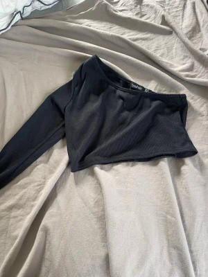 Svart oneshoulder croppad topp - Trendig svart oneshoulder topp från Boohoo. Croppad modell med lång ärm på ena sidan och ribbad struktur som ger en snygg och modern look. 