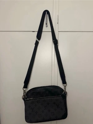 Svart axelväska med monogram - Snygg svart axelväska med justerbar axelrem och monogrammönster över hela väskan. Väskan har silverfärgade detaljer och dragkedja upptill. Perfekt för dig som vill ha en stilren och trendig accessoar.