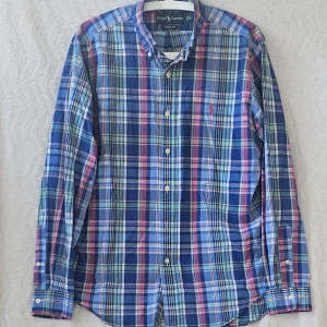 Rutig skjorta från Ralph Lauren M - Säljer en klassisk rutig skjorta från Ralph Lauren i storlek M. Skjortan har blå, röda, gröna och vita toner, lång ärm och knappar framtill. Tillverkad i mjuk bomull och har den ikoniska loggan broderad på bröstet. Perfekt för en stilren och avslappnad look.
