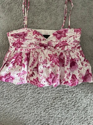Blommig linnetopp från Zara - Söt linnetopp från Zara med rosa blommönster på vit botten. Toppen har tunna axelband, v-ringning och en liten rosett framtill. Perfekt för sommarens varma dagar och ger en fräsch och trendig look. 