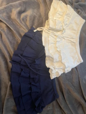 Två volangkjolar i blått och vitt. Den mörkblåa kjolen är i storlek xxs och den vita xs, men sitter ungefär likadant  - Säljer två korta kjolar med volanger, en i marinblått och en i vitt. Båda har resår i midjan och knytband framtill för en bekväm passform. Kjolarna är perfekta för sommarens alla tillfällen och har en mjuk känsla i bomull.