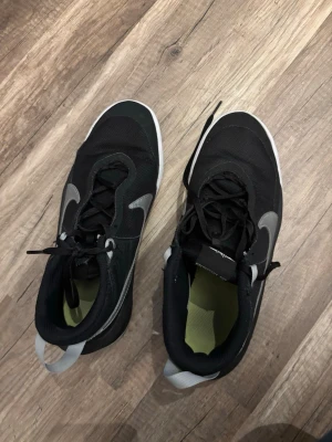 Svarta sneakers med grå detalj - Snygga svarta sneakers med stora grå swoosh-detaljer på sidorna och vit sula. Ovandelen är i mesh och syntetmaterial för bra andningsförmåga. Skorna har svarta skosnören och en vit hälflik för enkel påtagning. Perfekta för en sportig och trendig look.