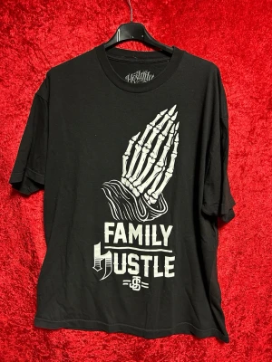 Svart t-shirt Family Hustle Hostility XL - Cool svart t-shirt från Hostility med tryck av skelett-händer i bön och texten 'Family Hustle' på framsidan. Tillverkad i 100% bomull och har en klassisk passform med korta ärmar och rund hals. Perfekt för dig som gillar streetwear och statement-plagg. Använd några gånger. Storlek XL.